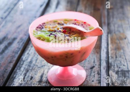 Cendol, ein malaysisches Dessert, das in einer roten Schüssel mit zerstoßenem Eis, grünem Gelee, roten Perlen, Kokosmilch und klebrigem Reis auf einem rustikalen Holztisch serviert wird. Stockfoto