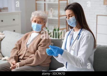 Ältere Frau in medizinischer Maske und weibliche Ärztin mit Spritze sitzend auf Sofa im Wohnzimmer Stockfoto