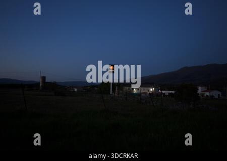Redaktionelles Bild des Schilds der Repsol-Tankstelle, beleuchtet bei Dämmerung im ländlichen Spanien mit kleinen Dorfgebäuden und Bergen im Hintergrund, die alles zeigen Stockfoto