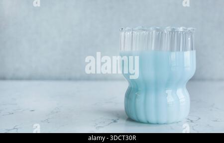 Minimalistisches ästhetisches Foto mit strukturiertem geripptem Glas, gefüllt mit einem cremigen, pastellblauen Getränk. Das Getränk, das Heidelbeermilch ähnelt, Stockfoto