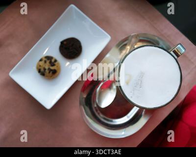 Flaches Foto mit einer Glasschale heißen Cappuccino oder Latte mit dickem, cremigem Milchschaum. Serviert auf glänzendem silbernen ovalen Tablett mit Teaspo Stockfoto