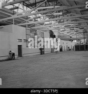 Slawjansk, Region Donezk, Ukraine, UdSSR. 1970er Jahre Ein authentisches historisches Schwarzweißfoto, das das riesige Innere der AIZ-Werkstätte zeigt. Hohe Decken sind mit Metallträgern und Oberlichtern ausgestattet. An der Wand hängt ein großes sowjetisches Propagandaplakat, das „50 Jahre Oktober“ mit Lenins Profil feiert, das vom Jubiläum übrig geblieben ist. Ein Arbeiter trinkt aus einem Brunnen auf der linken Seite, während andere in der Ferne laufen. Dieses Archivbild erfasst die Größe der Schwerindustrie und Ideologie. Eine friedliche Szene aus der Donbass-Vergangenheit. Stockfoto