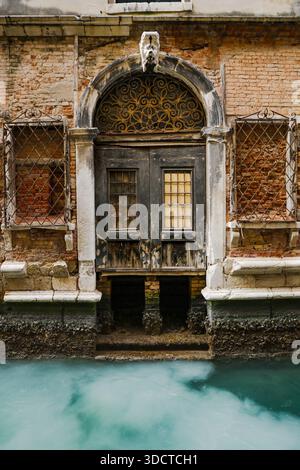Eine alte dunkle Holztür mit einem dekorativen bogenförmigen Heckspiegel, in eine verwitterte Ziegelmauer direkt über dem grünen Wasser eines Kanals von Venedig gesetzt. Stockfoto