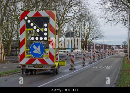 Großer mobiler LED-Trailer mit Verkehrspfeilen auf nasser Straße mit Kegeln, die den Verkehr bei Straßeninstandhaltungsarbeiten in den Niederlanden lenken. Stockfoto