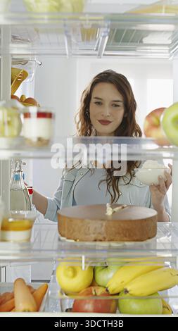 Glückliche, lächelnde Frau öffnet den Kühlschrank und schmeckt Schokoladenkuchen und Sahne. Sie zeigt Freude am Geschmack und genießt sichtbar die Leckerei. POV-Aufnahme von Stockfoto