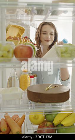 Schöne junge Frau öffnet die Kühlschranktür, nimmt eine Milchflasche heraus und bereitet gesundes Frühstück mit frischen Lebensmitteln zu. POV-POV-Aufnahme von Stockfoto