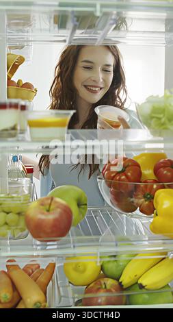 Die schöne Frau öffnet die Kühlschranktür, schaut hinein und nimmt einen süßen Snack raus. Sie bereitet eine gesunde Mahlzeit mit frischen Lebensmitteln zu. POV-Aufnahme von Stockfoto