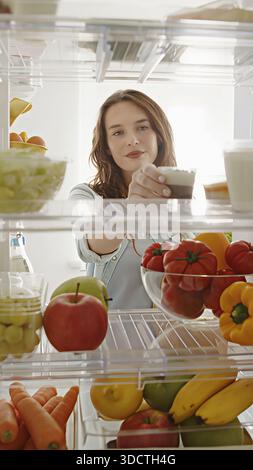 Die schöne Frau öffnet die Kühlschranktür, schaut hinein und nimmt einen süßen Snack raus. Sie bereitet eine gesunde Mahlzeit mit frischen Lebensmitteln zu. POV-Aufnahme von Stockfoto