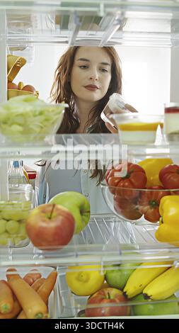 Die schöne Frau öffnet die Kühlschranktür, schaut hinein und nimmt einen süßen Snack raus. Sie bereitet eine gesunde Mahlzeit mit frischen Lebensmitteln zu. POV-Aufnahme von Stockfoto
