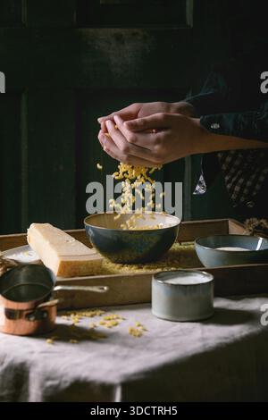 Junger Mann in Schwarz, der rohe ungekochte italienische Pasta-Coquillettes zum Kochen des Abendessens mit Parmesankäse, Sahne und Grießmehl auf Holztablett auf Küchen-Leinen-Tischtuch bestreut. Dunkler, rustikaler Stil. Stockfoto