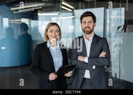 Zwei lächelnde Geschäftsleute, eine Frau mit einem Klemmbrett und ein Mann mit überkreuzten Armen, die selbstbewusst in einem modernen Glasbüro stehen und Teamarbeit und Erfolg darstellen Stockfoto