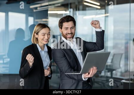 Professionelle Geschäftsleute feiern aktiv Erfolge und Erfolge, schauen mit einem Laptop in einer Büroumgebung zufrieden und demonstrieren Teamarbeit und Zusammenarbeit, nachdem sie gute Nachrichten erhalten haben Stockfoto