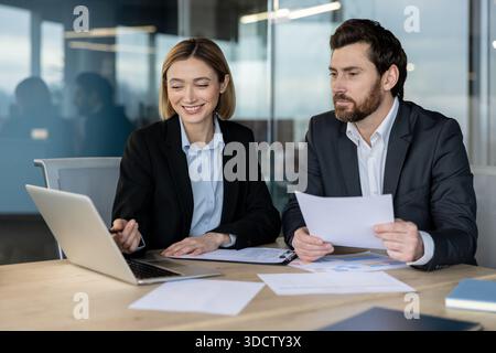 Zwei verschiedene Geschäftsleute arbeiten in einem modernen Büro zusammen, eine Frau, die auf einen Laptop-Bildschirm zeigt, während ein Mann Dokumente in der Hand hält, Projektdetails und Strategie diskutiert Stockfoto