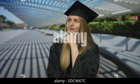 Eine junge Frau in Abschlusskleidung steht nachdenklich in einer Stadtstraße unter moderner Architektur und denkt über ihre Zukunft nach. Stockfoto