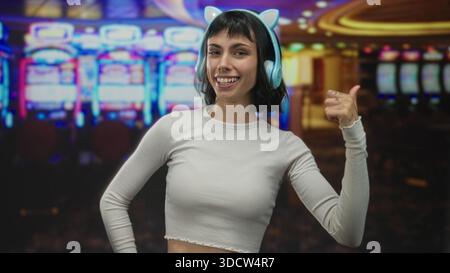 Frau mit Kopfhörern zeigt den Daumen auf Spielautomaten in einem von Neonlichtern beleuchteten Kasino-Gebäude; Glücksspiel-Risiko erregt die Spannung. Stockfoto
