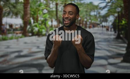 Junger afroamerikanischer Mann mit geballten Fäusten und sichtbaren Zahnspangen lächelnd auf der belebten Strandpromenade mit Palmen; Triumph. Stockfoto