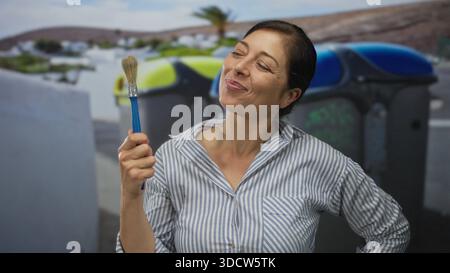 hispanische Frau mittleren Alters hält Pinsel in der Hand, indem sie Abfalleimer auf der Straße recycelt; Kreativität Verspieltheit Zufriedenheit Kunstfertigkeit. Stockfoto