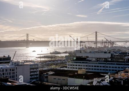 Lissabon Portugal 06 Dezember 2025 Niedrige Sonne reflektiert den Tejo mit einer Hängebrücke und Containerhafenkränen Stockfoto