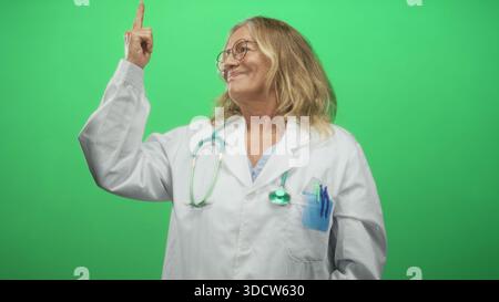 Seniorärztin mit Stethoskop und Stiften in der Tasche zeigt Zeigefinger nach oben zum grünen Bildschirm im Atelier; Vertrauensanleitung. Stockfoto