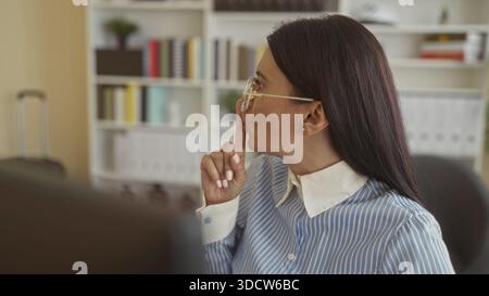 Frau in Brille, die auf einen Monitor zeigt und den Finger auf die Lippen legt, während sie mit der Hand in einem mit Büchern gefüllten Büro des Gebäudes Gesten macht; konzentriere dich Stockfoto