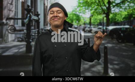 Mann Koch in schwarzer Uniform, der Koch Kuss Geste auf einer Stadtstraße mit Gesicht und Hand macht; stolz Handwerkskunst Gastfreundschaft. Stockfoto