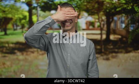 Der kaukasische Mann im Park bedeckt seine Augen verspielt, lächelt, während er draußen in lässigem Hemd steht, umgeben von hellen Bäumen unter einem sonnigen Himmel, überträgt Stockfoto