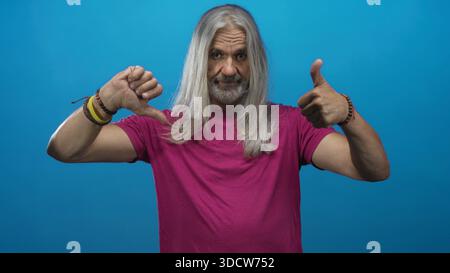 Mann mittleren Alters mit langen grauen Haaren, die Daumen nach oben und nach unten zeigen, während er ein magentafarbenes Hemd in einem blauen Studio trägt; Ambivalenz. Stockfoto