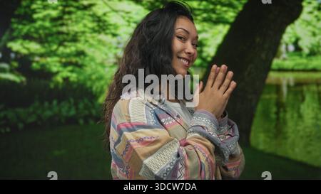 Frau mit Palmen zusammengedrückt nahe der Brust im grünen Park bei einem Baum, freundlich lächelnd und den Betrachter anschauend; Freude Dankbarkeit. Stockfoto
