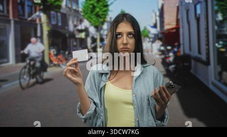 Junge hispanische Frau mit Kreditkarte und Smartphone auf einer Stadtstraße, rollende Augen und zuckte mit der Hand nach oben; Frustration. Stockfoto