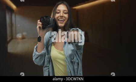 Junge hispanische Frau lächelt und hält die Kamera mit der Objektivkappe in der Hand, zeigt mit dem Finger auf das Objektiv im Studio; spielerische Verbindung. Stockfoto