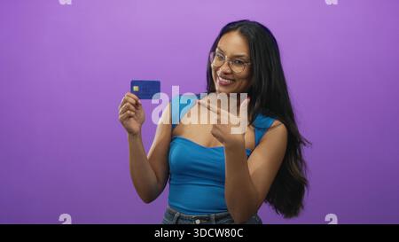Frau, die eine Kreditkarte hält und mit dem Finger auf die Karte zeigt, im violetten Hintergrund des Studios, zeigt Chip und lächelnd; finanzielles Vertrauen. Stockfoto