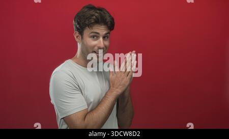 Junger hispanischer Mann lächelt und klatscht in einem roten Studio in hellgrauem T-Shirt und blickt in Richtung Kamera; fröhliche Wärme. Stockfoto