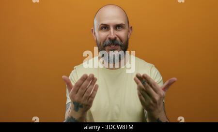 Ein kahlköpfiger Mann mit Händen gegen die orangefarbene Wand, zeigt Tattoos und einladenden Ausdruck, trägt ein gelbes Hemd in einem isolierten Studio-Set Stockfoto