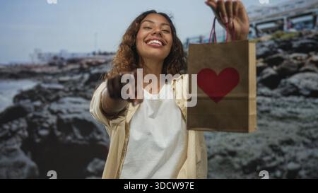 Frau zeigt mit dem Finger auf die Kamera, während sie eine Papiertüte mit rotem Herzen an der Küste hält; Freude schenken. Stockfoto