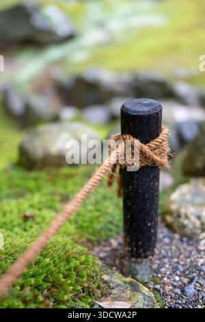 Ein dickes Seil ist um einen dunklen Holzpfosten gebunden, der einen Weg durch einen moosigen Garten mit Felsen und Kieselsteinen markiert. Stockfoto
