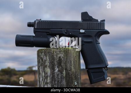 Moderne taktische, halbautomatische Pistole mit rotem Punkt und Taschenlampe auf einem Holzpfosten vor bewölktem Himmel. Stockfoto