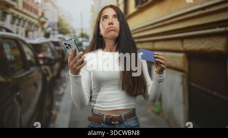 Frau mit Smartphone und Kreditkarte auf der Stadtstraße mit geparkten Autos und Gebäudefassade; frustriertes Zahlungsproblem. Stockfoto