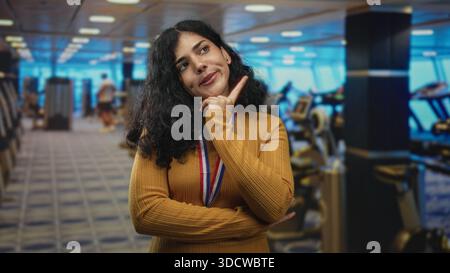 Arabische Frau mit Goldmedaille und Senfpullover zeigt den Finger in Richtung Kamera im Fitnessgebäude mit Laufbändern hinter sich, während sie lächelt; VI Stockfoto