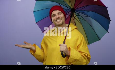 Junger Mann in gelbem Regenmantel hält einen bunten Regenschirm vor isoliertem lila Hintergrund, der die Palme mit einem selbstbewussten, schönen Ausdruck und Saibling ausdehnt Stockfoto