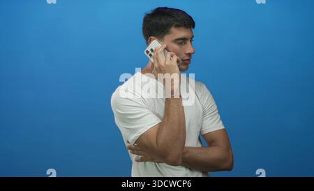 Junger Mann, der das Smartphone mit gekreuzten Armen in einem hellblauen Studio lässig am Ohr hält; konzentrierte Aufmerksamkeit. Stockfoto