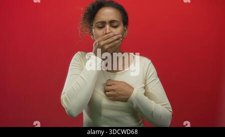 Frau, die den Mund mit der Hand bedeckt, Brust umklammert und Grimassen beim Husten im roten Studio-Set; Übelkeit. Stockfoto