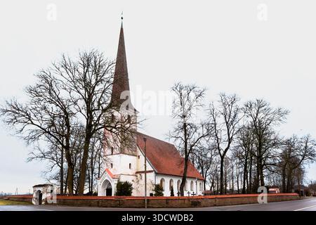 Außenansicht der lutherischen Nurme-Kirche in Lettland. Historisches weißes Gebäude mit einem hohen Turm, rotem Dach und einer Steinmauer an einem bewölkten Tag. Stockfoto
