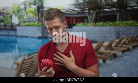 Mann mit rotem Apfel und berührender Brust mit rechter Hand im Hotelresort am Pool, rotes Poloshirt, neutraler Ausdruck, Gelassenheit. Stockfoto