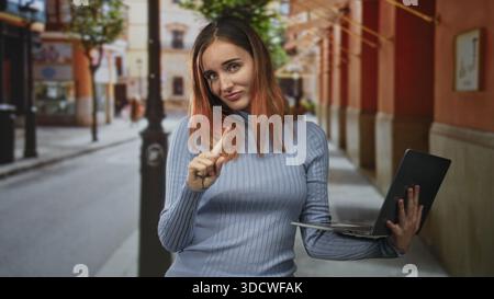 Frau, die einen Laptop hält, zeigt mit dem Zeigefinger auf eine Fußgängerzone, die von Bögen und Geschäften gesäumt ist, rote Haare und lässiger Pullover sichtbar; Selbstvertrauen. Stockfoto