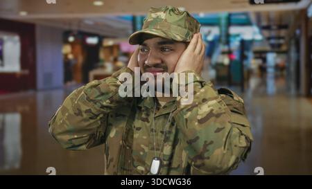 Hispanischer Mann in Tarnuniform bedeckt die Ohren mit beiden Händen inmitten des überfüllten Einkaufszentrums; Unwohlsein. Stockfoto