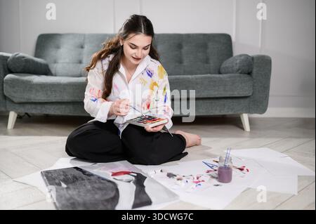 Junge europäische Frau in lackiertem Hemd sitzt zu Hause auf dem Boden und malte mit Aquarellen, mit Palette, Pinsel, Leinwand, lächelnd, mischen von Farben. Entspannte, kreative Atmosphäre im Wohnzimmer oder Studio Stockfoto