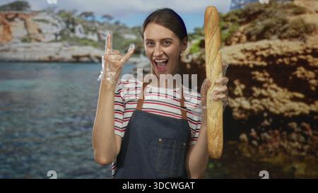 Frau mit Schürze und Kunststoffhandschuhen, die ein langes Baguette hält und im Studio Geste mit Steinhörnern zeigt; Freude. Stockfoto