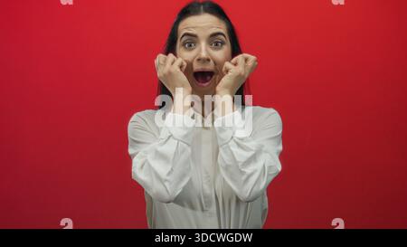 Frau, die ihre Begeisterung mit den Händen nahe dem Gesicht vor einer leuchtend roten Wand ausdrückt, intensive Emotionen und Energie vermittelt, in einem weißen Hemd steht, Captur Stockfoto