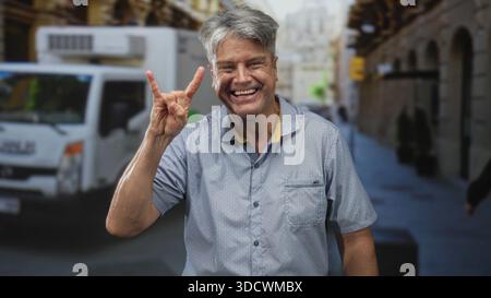 Mann lächelt und macht Steinhörner Hand Gesten, während er in einer überfüllten Straße mit Motorrollern und Lieferwagen steht; freudige Reise. Stockfoto