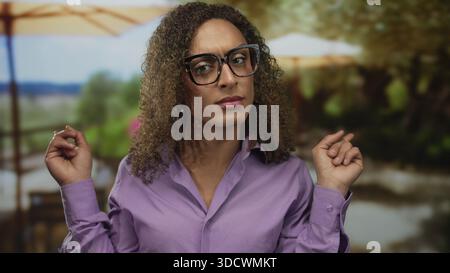 Frau mit lockigem Haar und Brille zeigt beide Zeigefinger seitwärts im Park mit Regenschirmen und Laub; Unentschlossenheit. Stockfoto